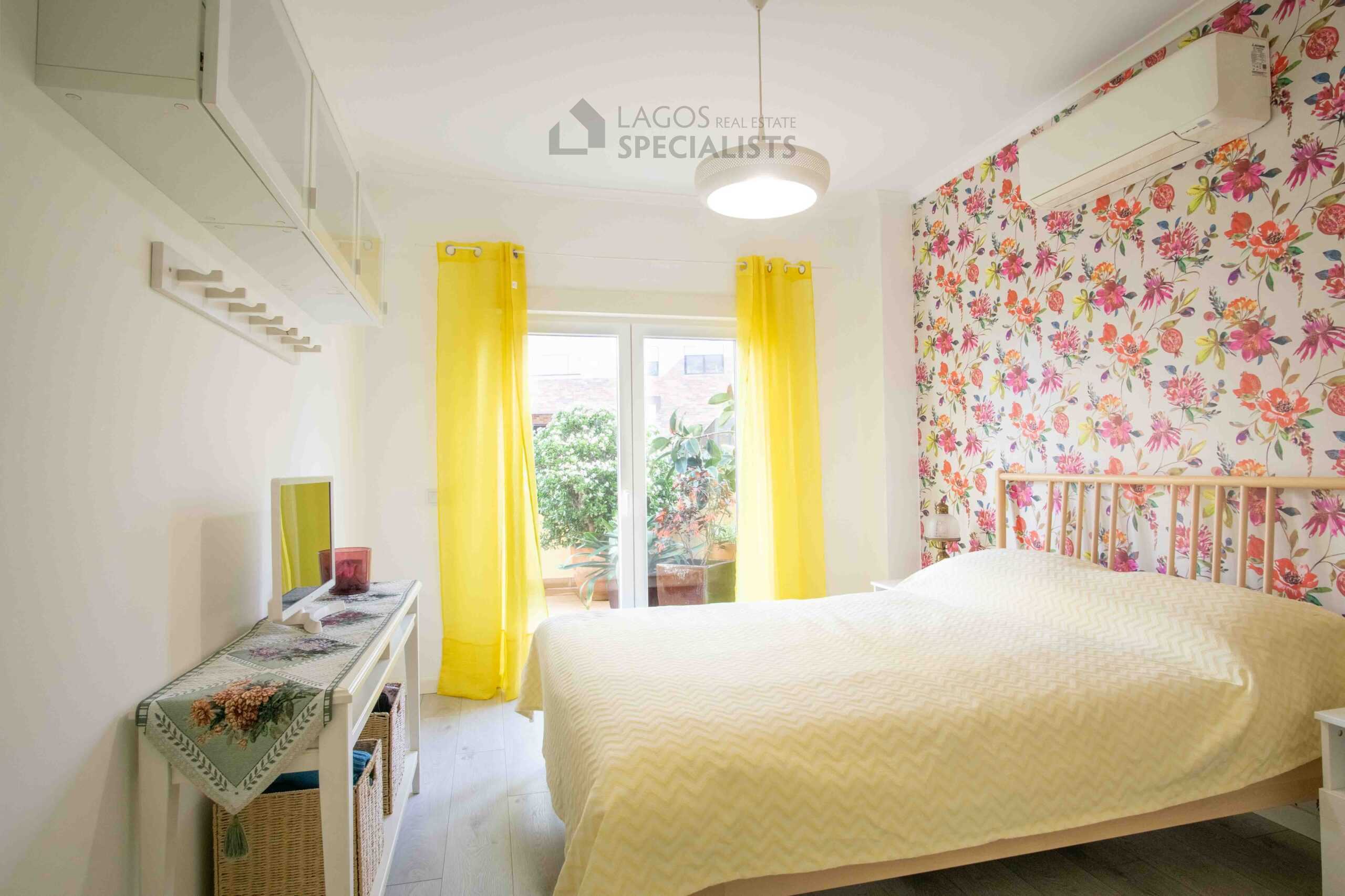 Light-filled bedroom with yellow curtains, floral feature wall, air conditioning, and direct access to a private balcony in Lagos apartment